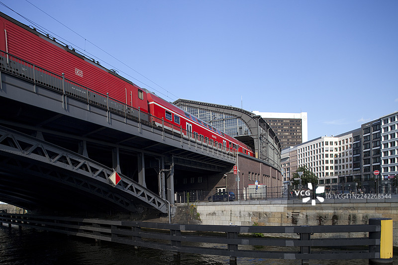 一辆红色火车正在柏林斯普雷河上的桥梁上行驶，背景是城市建筑，天空晴朗湛蓝，地点为德国柏林。图片素材