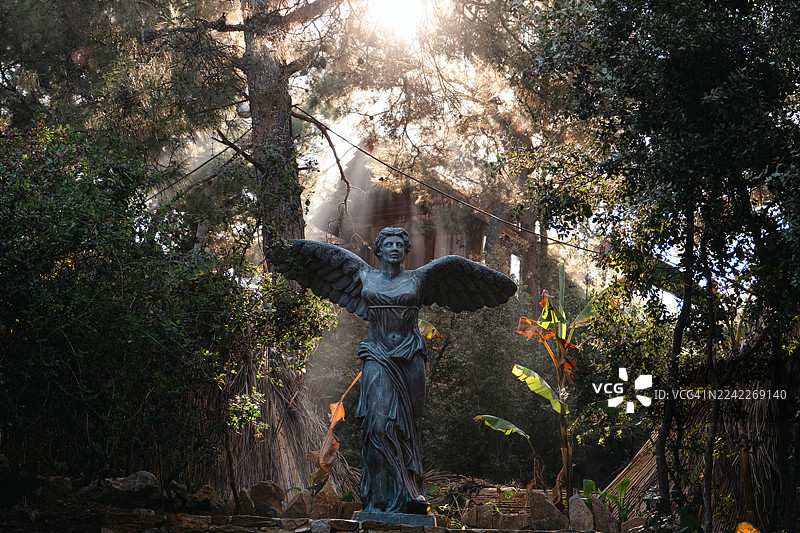 长着翅膀的雕像矗立在树林中，阳光透过树叶洒落图片素材