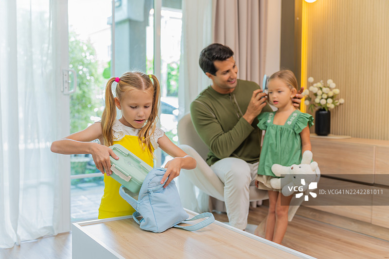 一家温馨的家庭住宅内，父亲正在帮女儿梳理头发，旁边还有一个孩子在收拾东西。图片素材