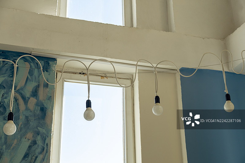 一串灯泡挂在白色的线上，悬挂在一扇明亮的窗户前，窗户后面是蓝色的墙。图片素材