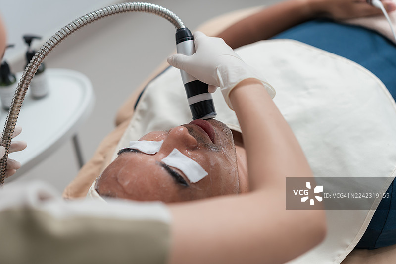 美容师为近距离拍摄的亚洲男性进行面部水疗护理，使用面部仪器和精华液。主打抗衰老、胶原蛋白再生和皮肤焕新。图片素材