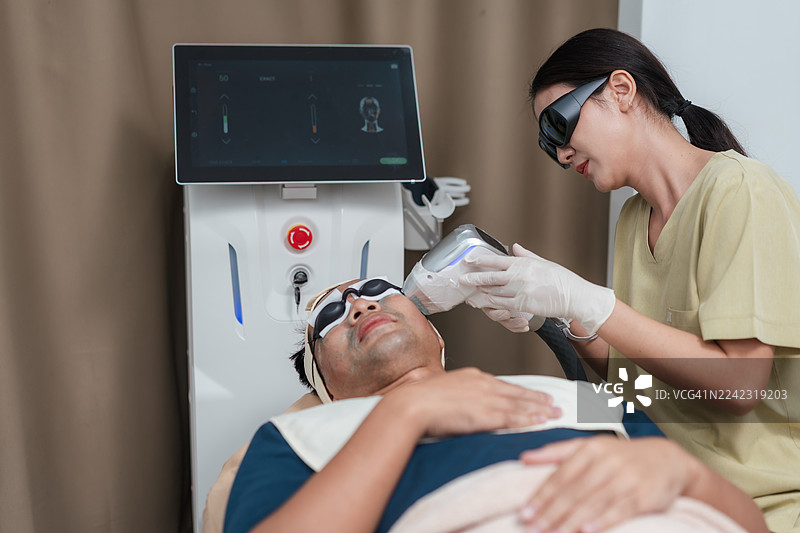 亚洲女美容师使用专业激光设备为男性患者进行治疗。一名男子接受激光嫩肤以获得清透、光泽的肌肤。治疗痤疮、毛孔粗大，改善衰老迹象。图片素材