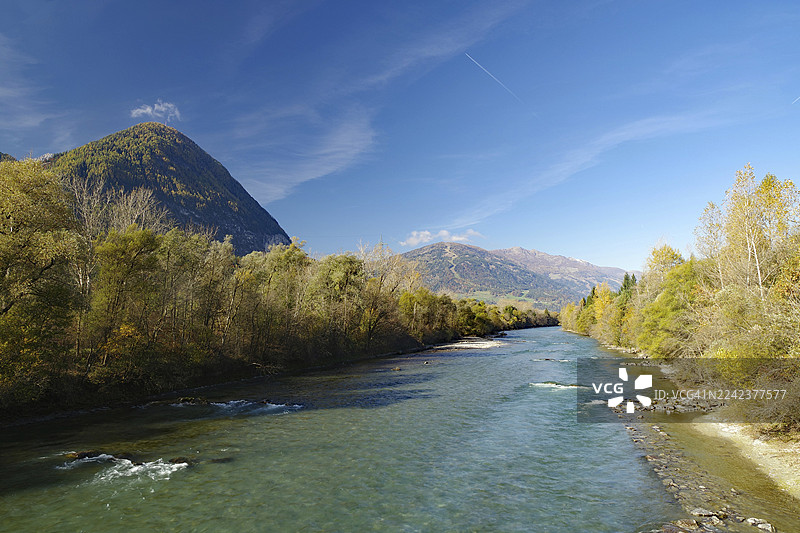 宽阔的河流蜿蜒穿过秋日的山谷，天空宁静，有细微的飞机尾迹，利恩茨，德拉瓦自行车道，卡林西亚，意大利图片素材