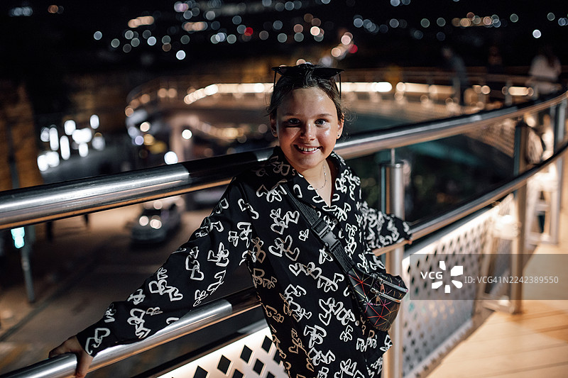 一位女士微笑着走在夜晚的城市步道上，背景是闪烁的灯光。图片素材