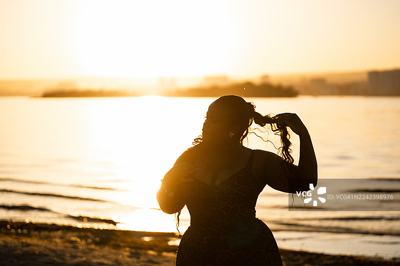 夕阳下，一位女士在海滩上，在温暖的金色光线下调整她的头发的剪影图片素材