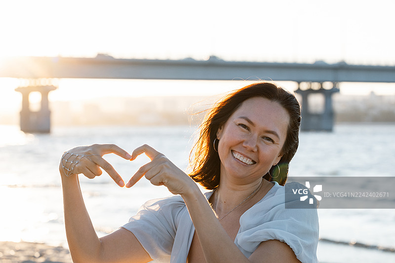 日落时分，女子在水边用双手比出心形，面带微笑图片素材