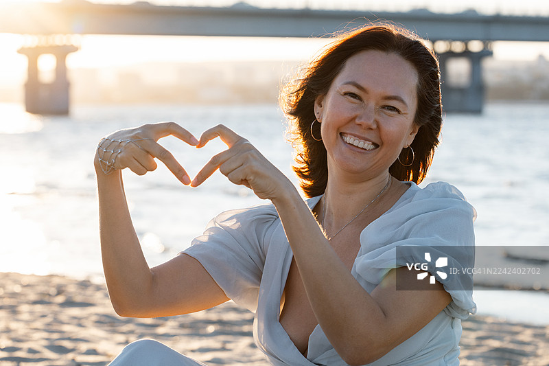 日落时分，女子在水边海滩上微笑，双手比出爱心形状图片素材