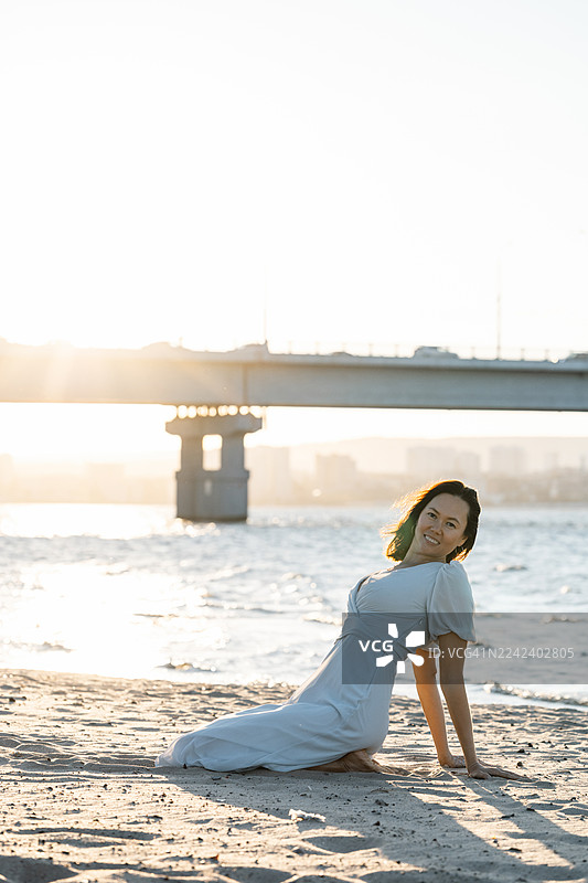 一位身穿飘逸白裙的女子在日落时分于桥边海滩放松图片素材