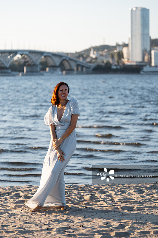 身穿浅蓝色连衣裙的女子在海滩上行走，背景是城市天际线和桥梁图片素材