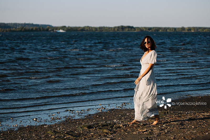 金色的日落时分，身穿白色连衣裙的女子赤脚走在湖边沙滩上，阳光照耀着岸边。图片素材