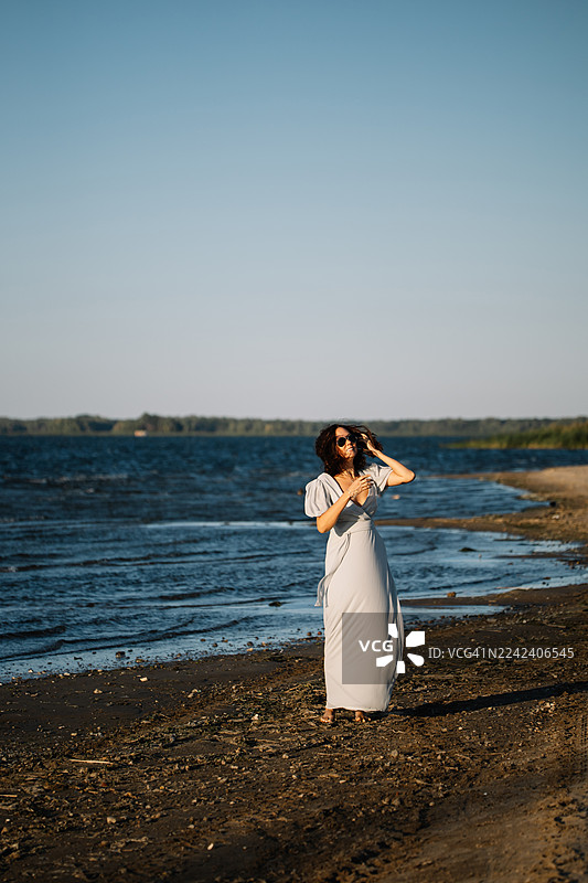 一位身着飘逸白裙、戴着太阳镜的优雅女士在阳光明媚的海滩上漫步，身后是海岸风光图片素材