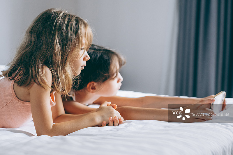 两个年轻女孩躺在床上，在自然光下放松地专注地看着智能手机上的内容。图片素材