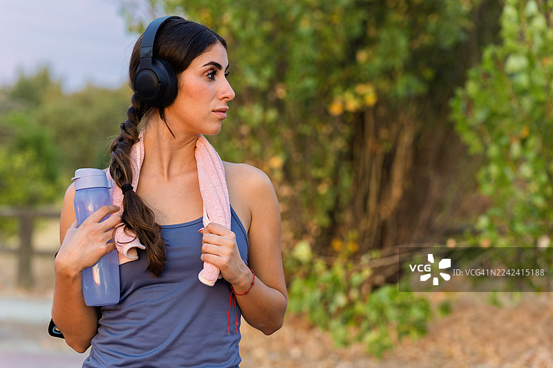 年轻女子戴着耳机，带着运动水壶在户外锻炼图片素材