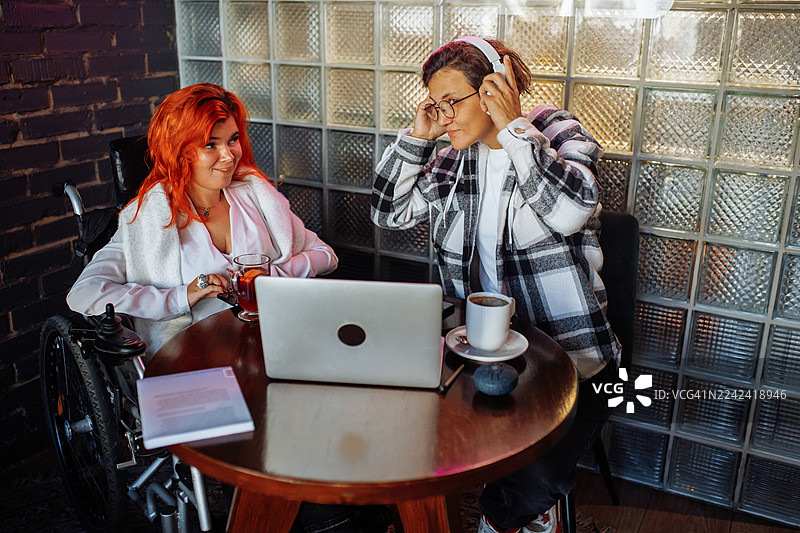 两位朋友在一家舒适的咖啡馆，围坐在圆桌旁，桌上放着笔记本电脑和饮品，分享着片刻的时光。图片素材