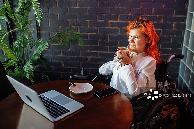 一名坐在轮椅上的女士在咖啡馆里，旁边放着笔记本电脑、手机和绿植，她正享受着咖啡。图片素材