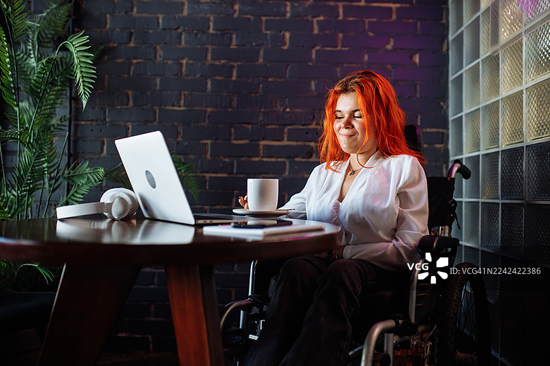 一位女士坐在轮椅上，在现代咖啡馆的办公桌前使用笔记本电脑，旁边放着一杯咖啡。图片素材