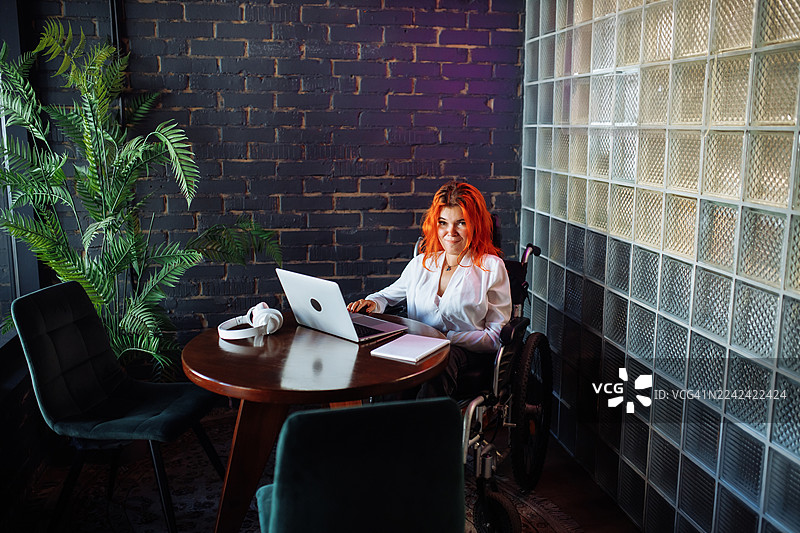 一位坐在轮椅上的女性在现代办公室的圆形木桌旁使用笔记本电脑，并戴着耳机工作图片素材