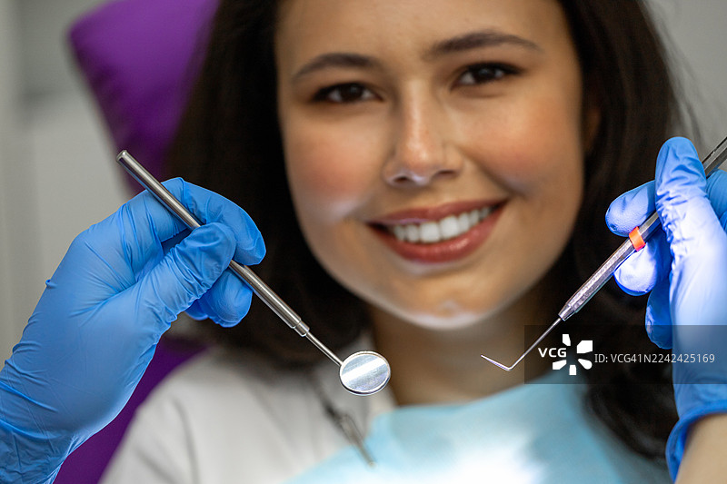一位戴着手套的手拿着两种牙科器械的患者，坐在牙科椅上，面带微笑。图片素材