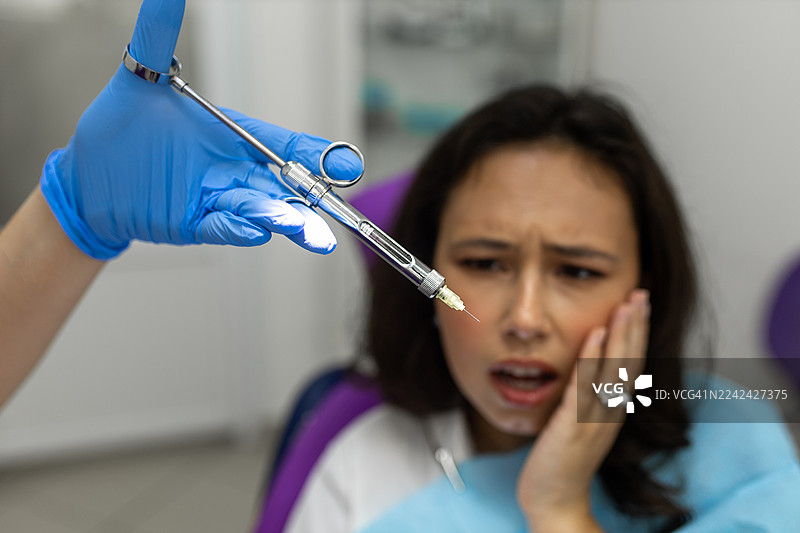 正在进行的牙科手术：诊所里，一位恐惧的患者和戴着手套、拿着注射器的手图片素材