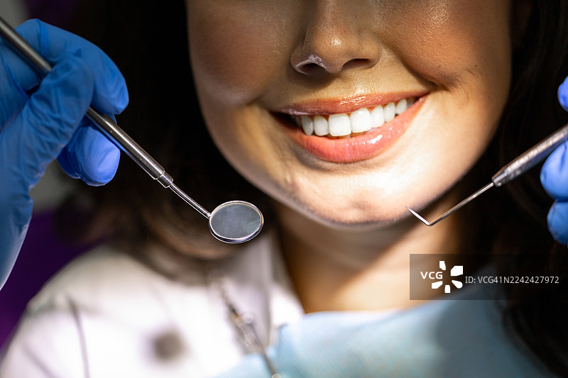 在临床牙科环境中，一名患者在牙科检查时对着镜头微笑，医生使用口腔镜和探针进行检查。图片素材
