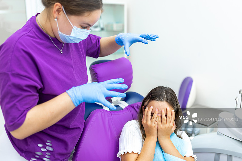 小儿牙科就诊，一名紧张的女孩坐在紫色椅子上，旁边是戴着口罩的牙医图片素材