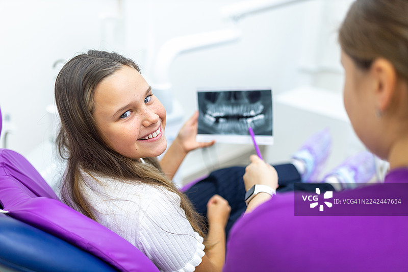 年轻女孩坐在牙医椅上，在儿科检查时，牙医向她解释牙科X光片，女孩面带微笑。图片素材