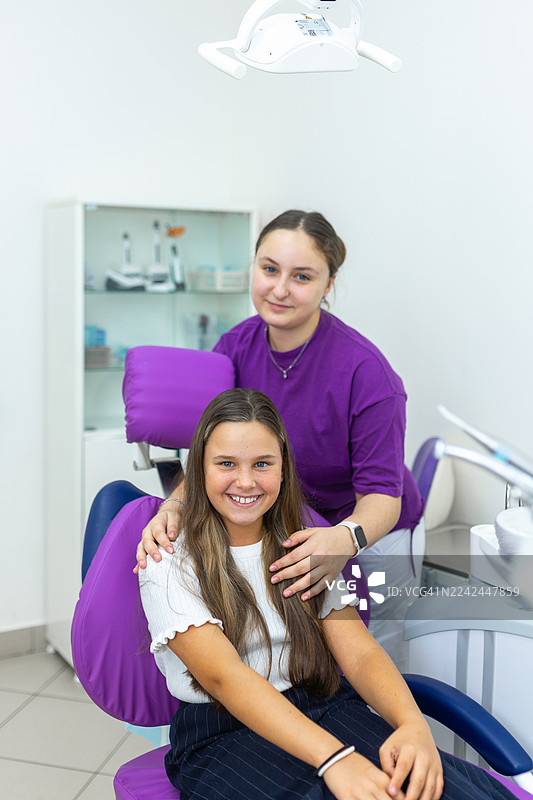 一位年轻的患者坐在紫色的牙科椅上，旁边有一位关怀备至的助理，在现代化的诊所里，患者开心地笑着。图片素材