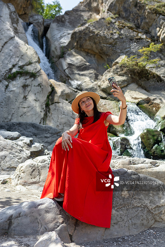 身穿红裙的女子在阳光明媚的户外，瀑布旁的岩石上自拍图片素材