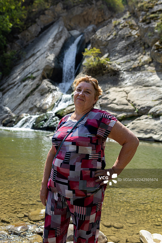 身穿几何图案粉色套装的女士自信地微笑着，在瀑布和岩石水池旁摆姿势图片素材
