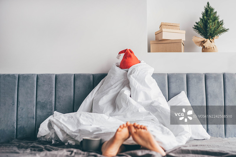 一个女人坐在床上，全身裹在白色羽绒被里，戴着圣诞帽，表情忧郁而厌倦。图片素材