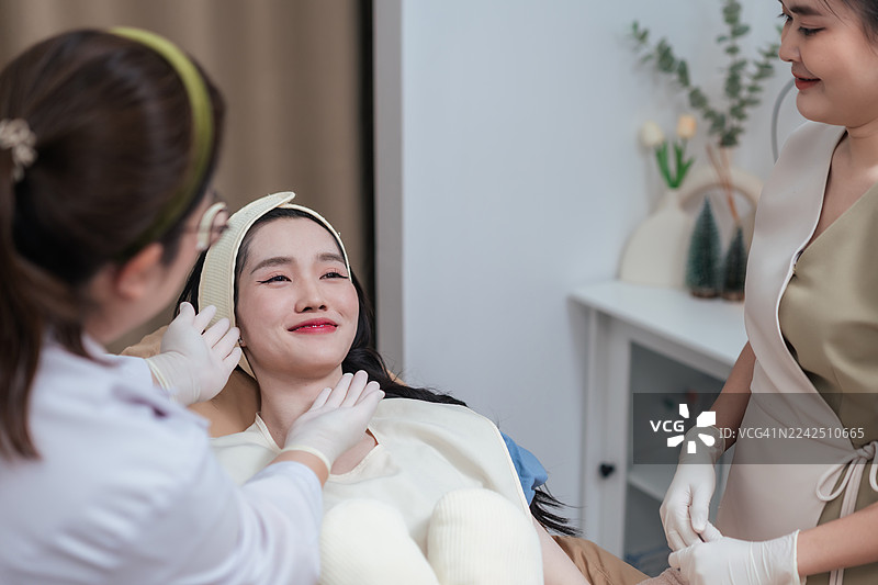 面部美容治疗前的快乐亚洲女性。皮肤科医生在现代美容诊所检查女性皮肤，准备进行美容治疗。图片素材