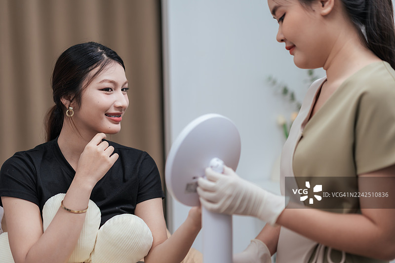 年轻的亚洲女性在美容诊所接受填充治疗后照镜子微笑。女性在填充治疗后感到自信。图片素材