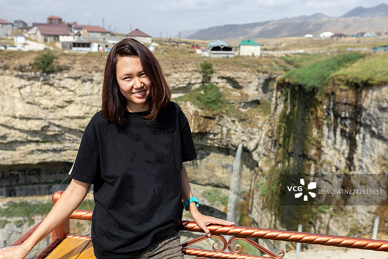 一位女士面带微笑地倚靠在栏杆上，背景是风景优美的峡谷观景点，可以看到瀑布和村庄。图片素材
