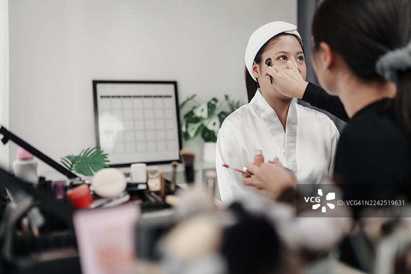 亚洲新娘在工作室由专业化妆师上底妆，化妆师用刷子为新娘脸部均匀涂抹粉底。图片素材