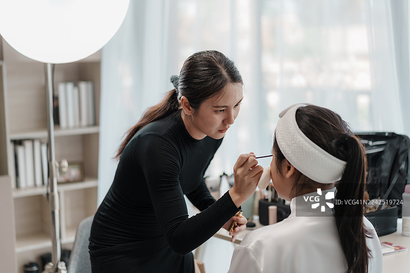 亚洲化妆师为新娘化妆，涂抹眼影和粉底。美容师专注于新娘妆容细节，展现美容行业的专业技能。图片素材