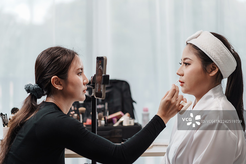 亚洲化妆师在工作室用唇刷精准地为新娘涂抹口红，并用智能手机录制口红过程。美容护理、专业知识和网红生活方式。图片素材
