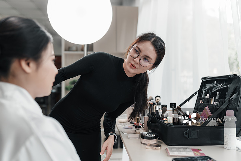 亚洲女性专业化妆师在工作室工作区检查新娘完成妆容后的细节。图片素材