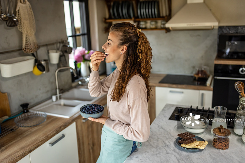一位女士在家中舒适的厨房里烘焙点心，同时享用着蓝莓。图片素材