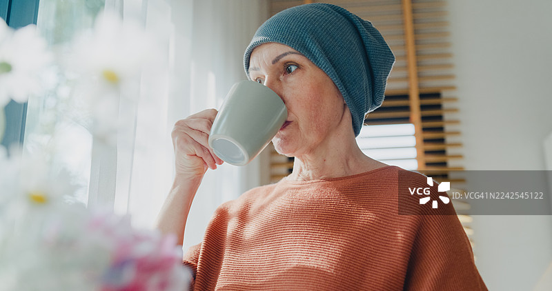 一位戴着头巾的年长女性癌症患者，在家中客厅的窗边站着喝茶。癌症生存概念。图片素材