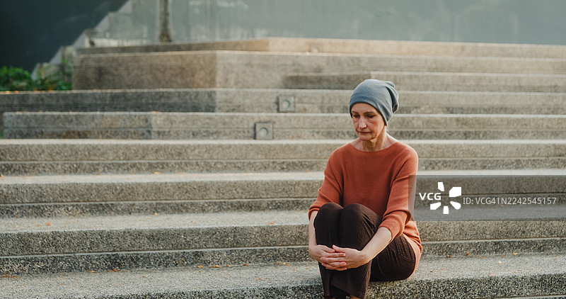 一位戴着头巾的年长女性癌症患者，在康复期间独自坐在公园里沉思。与癌症共存的概念。图片素材