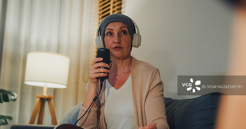 一位戴着头巾的年长女性癌症患者，在家里的客厅里，晚上戴着耳机，通过播客讲述她的癌症康复故事。与癌症共存的概念。图片素材