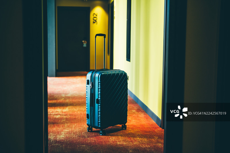 一个现代的深蓝色行李箱，位于一间黑暗、氛围感十足的酒店走廊中央，走廊墙壁是明亮的黄色，地毯是温暖的橙色。图片素材