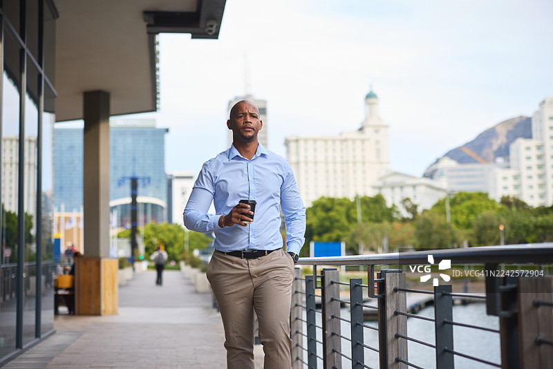 穿着蓝色衬衫的自信男人在城市滨水区行走，一手拿着咖啡，目光向前图片素材