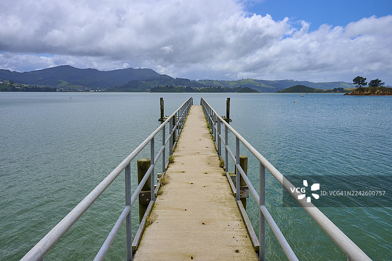 长长的栈桥延伸至平静的水面，背景是多云的天空和远处的山峦，地点位于新西兰南岛但尼丁奥塔哥的布罗德湾。图片素材