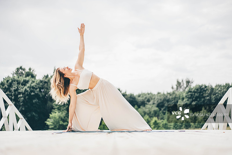 一位女子在绿意盎然的树林背景下，于一座白色木桥上摆出瑜伽姿势。开阔的空间和无垠的天空象征着个人成长、灵活性以及与大自然的连接。图片素材