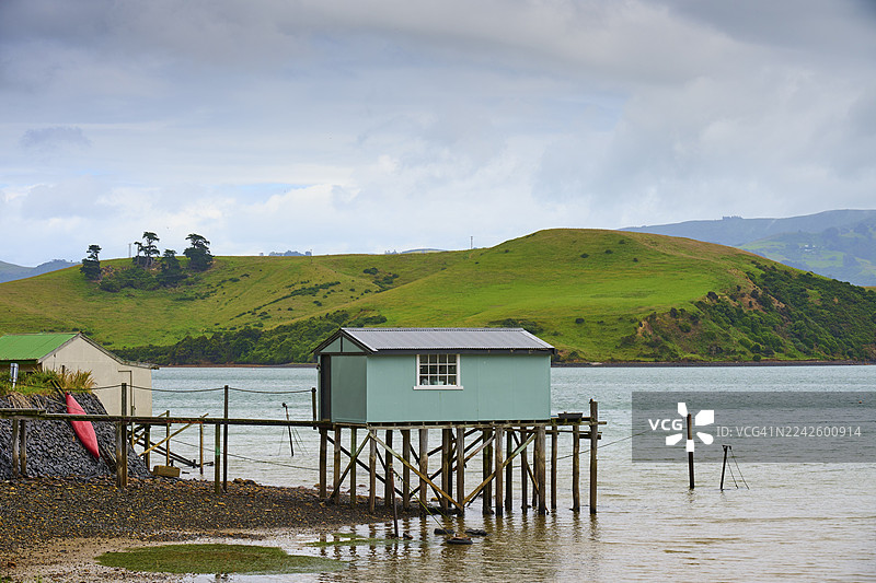 新西兰南岛但尼丁奥塔哥布罗德湾，建在水边高跷上的小屋，背景是青翠的山峦和多云的天空图片素材