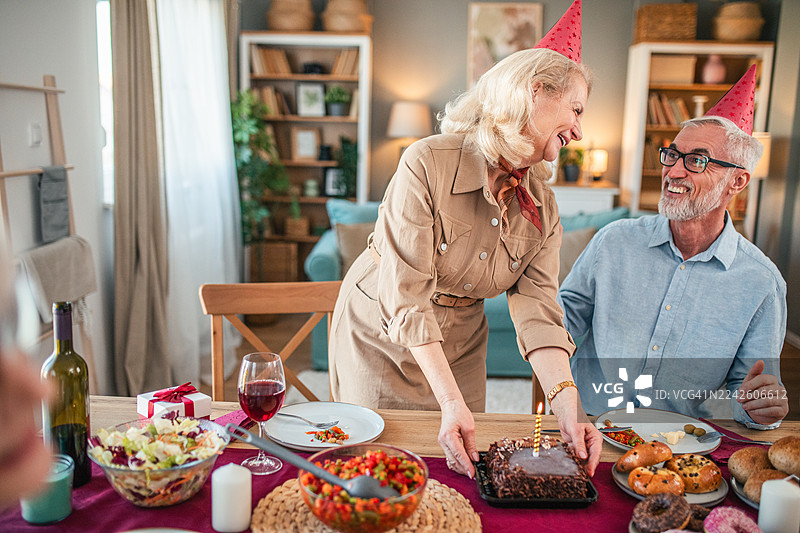 妻子为丈夫送上点燃蜡烛的蛋糕，他正与朋友们一同庆祝。大家戴着派对帽，享受着家中生日派对的欢乐气氛。图片素材