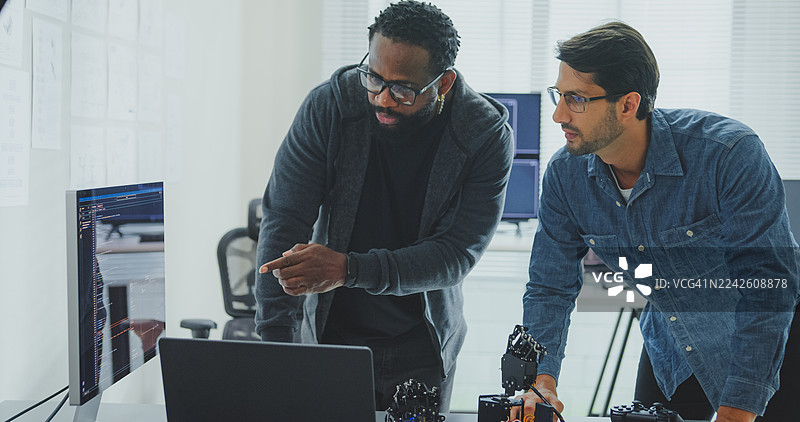 黑人和西班牙裔男性工程师程序员在科技办公室合作开发机器人手原型，使用计算机控制的机器人手臂电路。软件开发人员，人工智能。图片素材