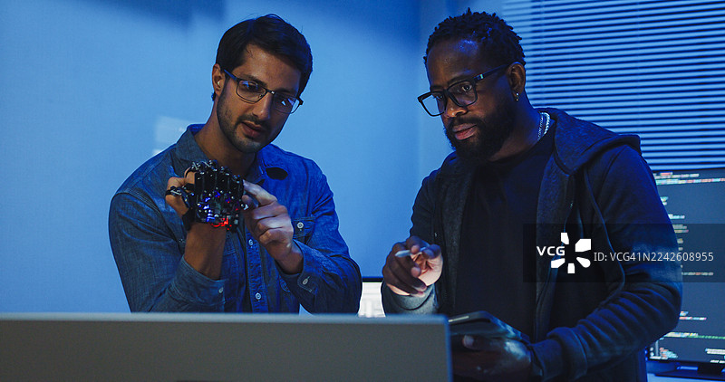 黑人和西班牙裔男性工程师程序员在科技办公室的夜晚一起开发机器人手原型，使用计算机控制的机械臂电路。软件开发人员，人工智能。图片素材