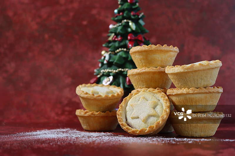 单个肉馅饼堆叠在肉馅饼塔上，顶部装饰着带有糖粉的星形酥皮盖，一堆节日甜点酥皮点心，圣诞树模型，斑驳的红色背景，焦点在前景图片素材